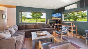 Ein modernes Wohnzimmer mit einer Eckcouch, einem Couchtisch mit Schachfiguren, einem Fernseher und einem Bücherregal. Große Fenster bieten Blick auf eine grüne Landschaft.