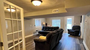 A spacious living room with a sectional sofa, a recliner, and a small table. There are windows providing natural light and a door leading to another room.