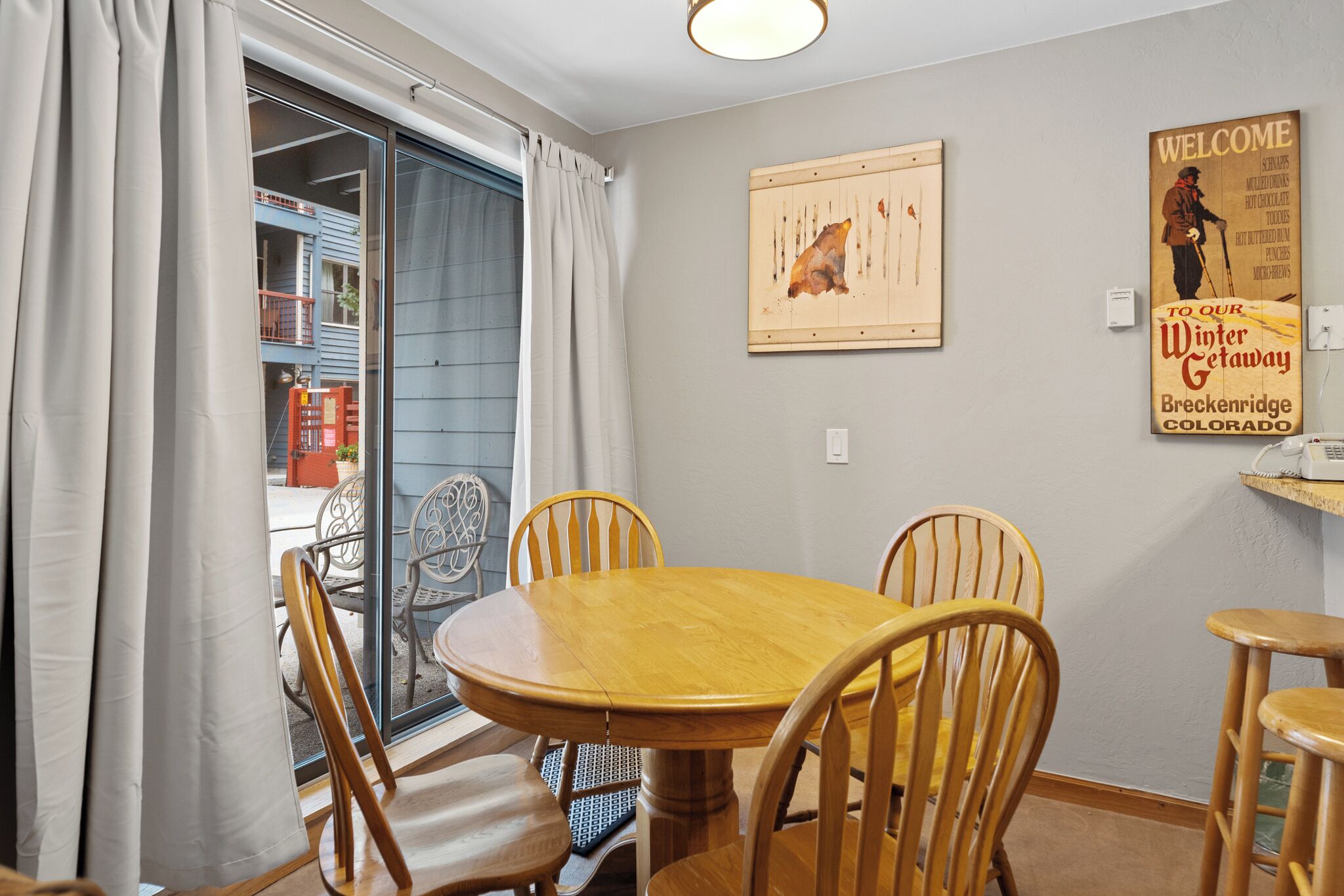 Un área de comedor con una mesa y sillas de madera, un puerta corredera de vidrio que da a un balcón exterior, y una pared con un letrero de bienvenida y un cuadro enmarcado.
