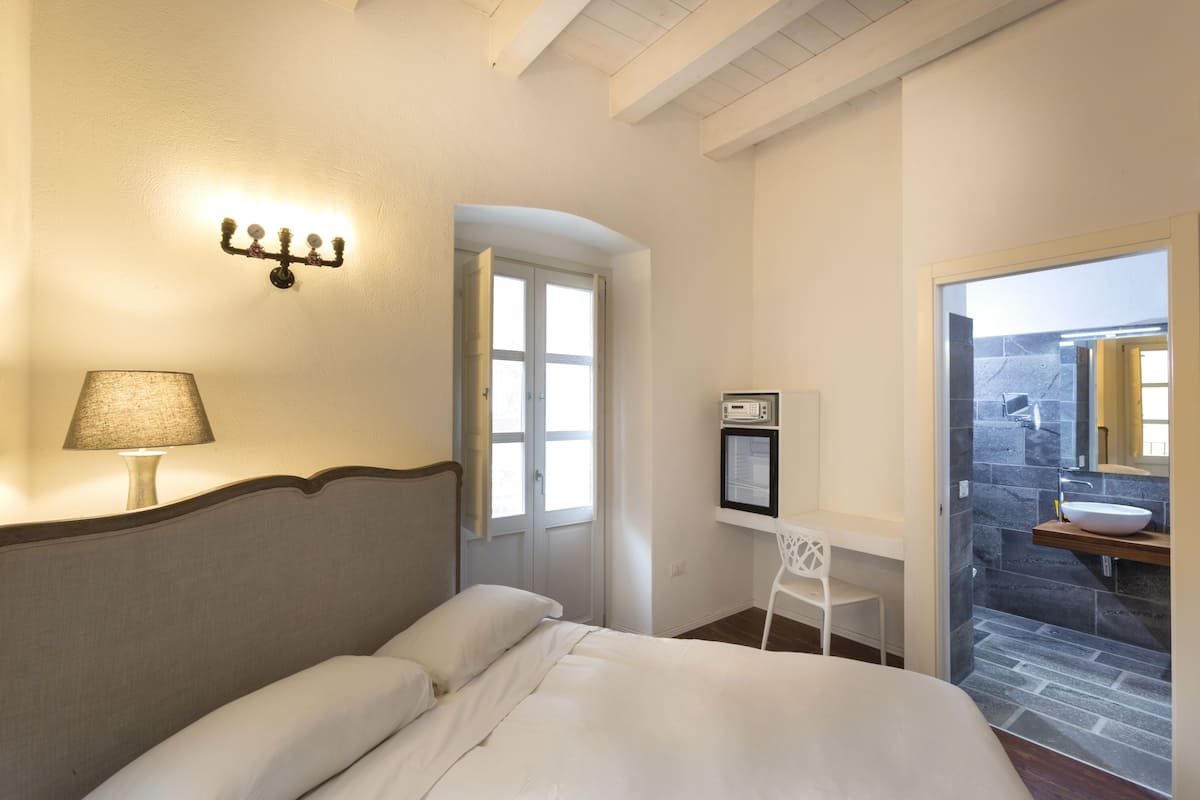 A hotel room with a bed, a desk with a lamp, a wall-mounted light fixture, a small white chair, and a bathroom visible through an open door.