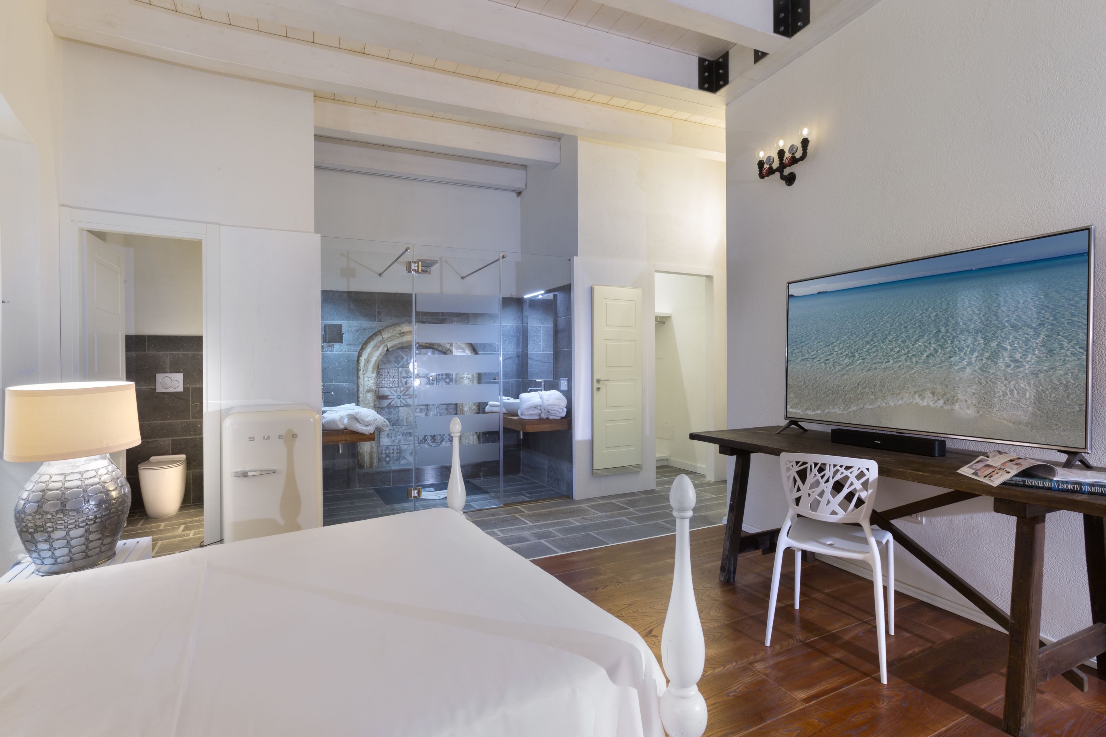 A hotel room with a bed, a desk with a TV, a chair, a lamp, and a shower area.