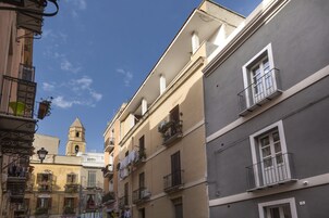 Una calle estrecha con edificios que tienen balcones y una torre de campanas al fondo.