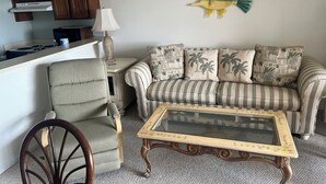 Un salon avec un canapé à rayures, une chaise en bois, une table basse en verre et un coin cuisine doté de meubles en bois et d’une cuisinière.