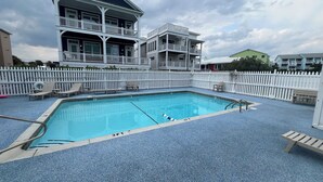 Une piscine rectangulaire bordée d’une terrasse bleue, entourée d’une clôture blanche en bois, et un bâtiment à deux étages avec des balcons en arrière-plan.