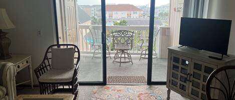 Una sala de estar con televisor, sofá, sillón y mesa. Hay un balcón con sillas y vistas a edificios.