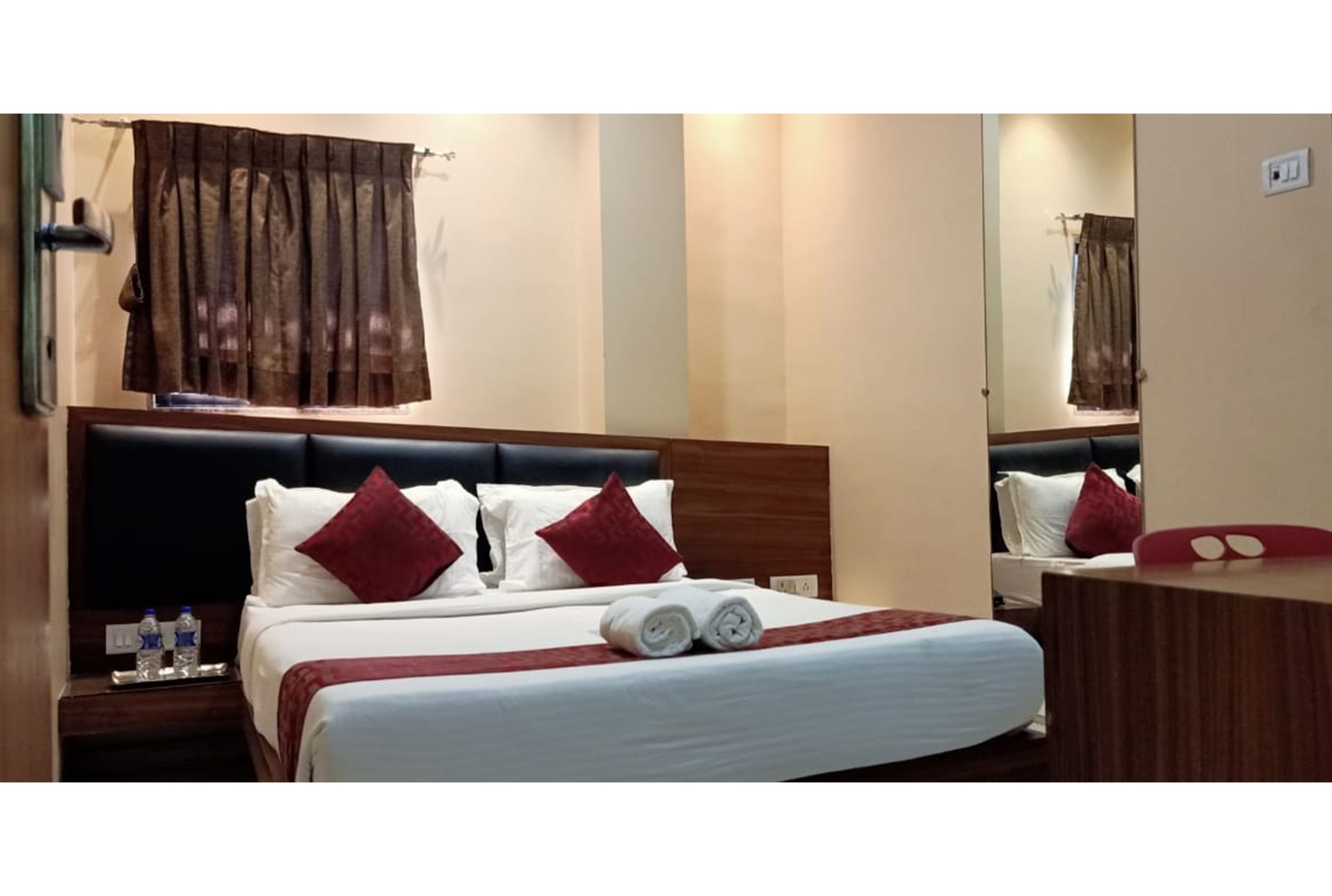 A hotel room with a bed, a nightstand with water bottles, a mirror, and a wooden dresser.