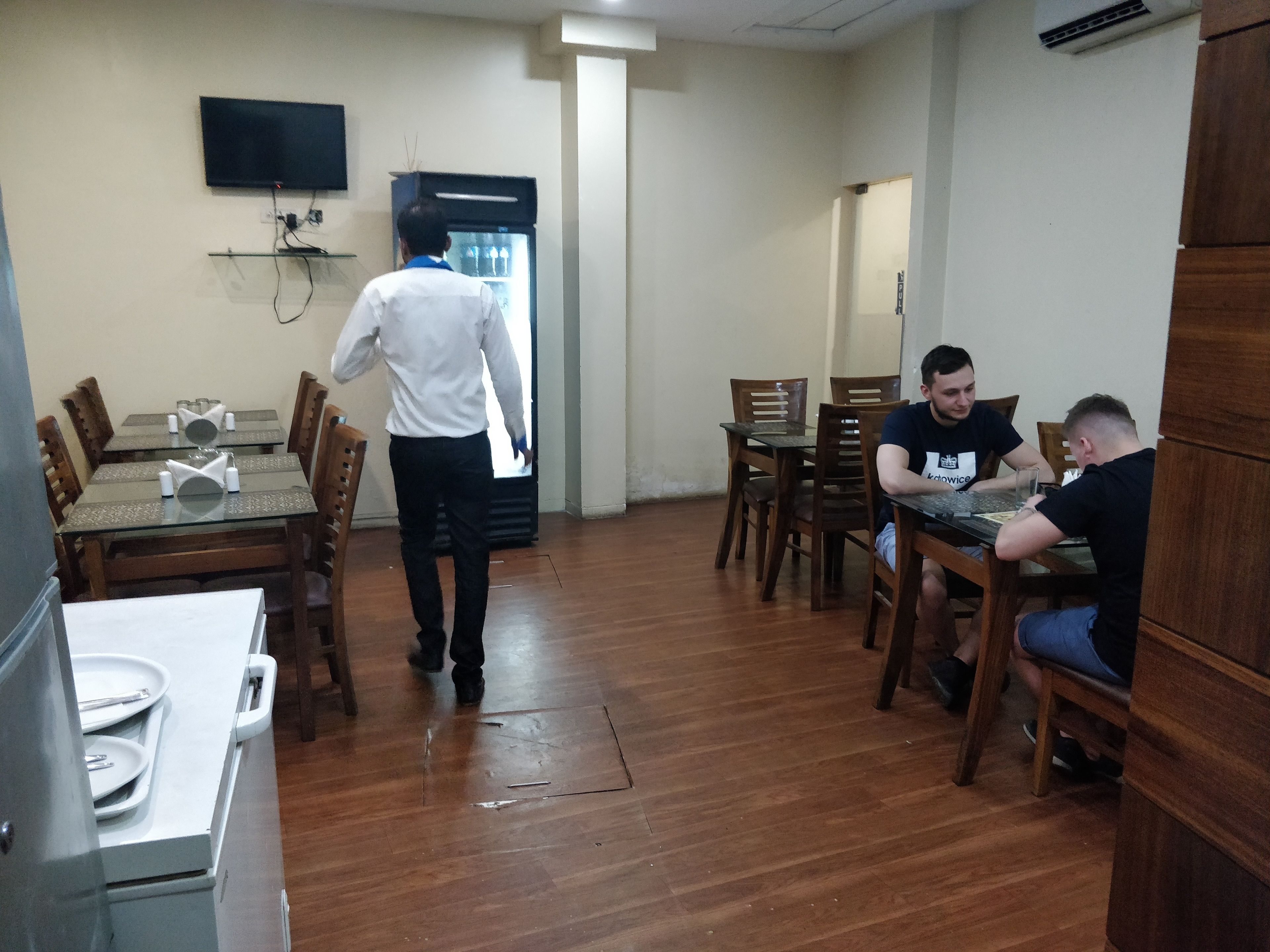 Un coin repas avec des tables et des chaises en bois, un réfrigérateur et un téléviseur à écran plat fixé au mur.