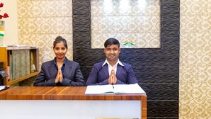 Two individuals seated at a wooden reception desk with a patterned wall behind them. - Hotel Apex International (Mumbai)