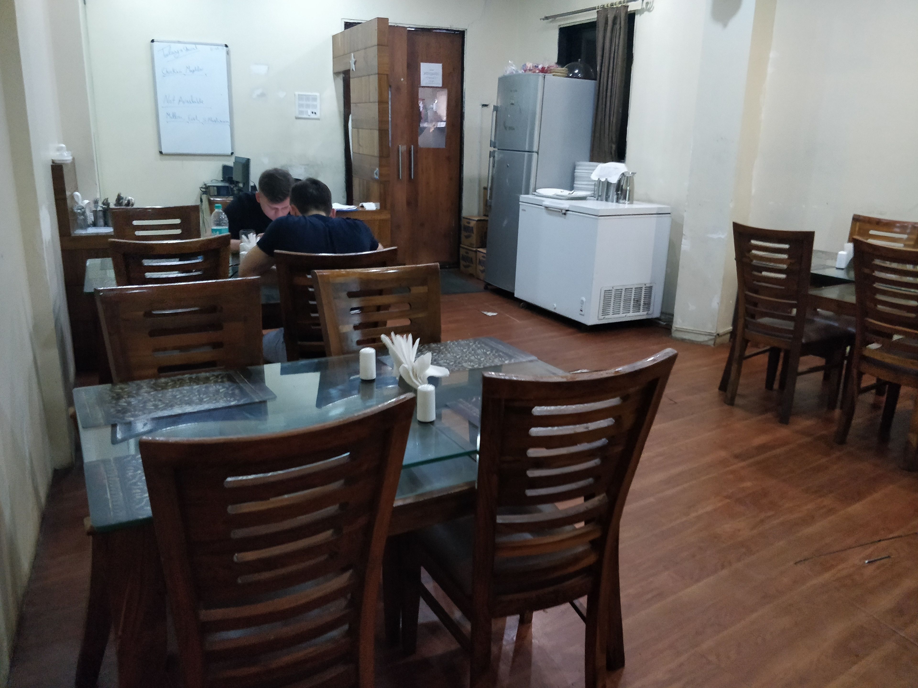 A dining area with wooden chairs and a glass table, a refrigerator, and a whiteboard on the wall.