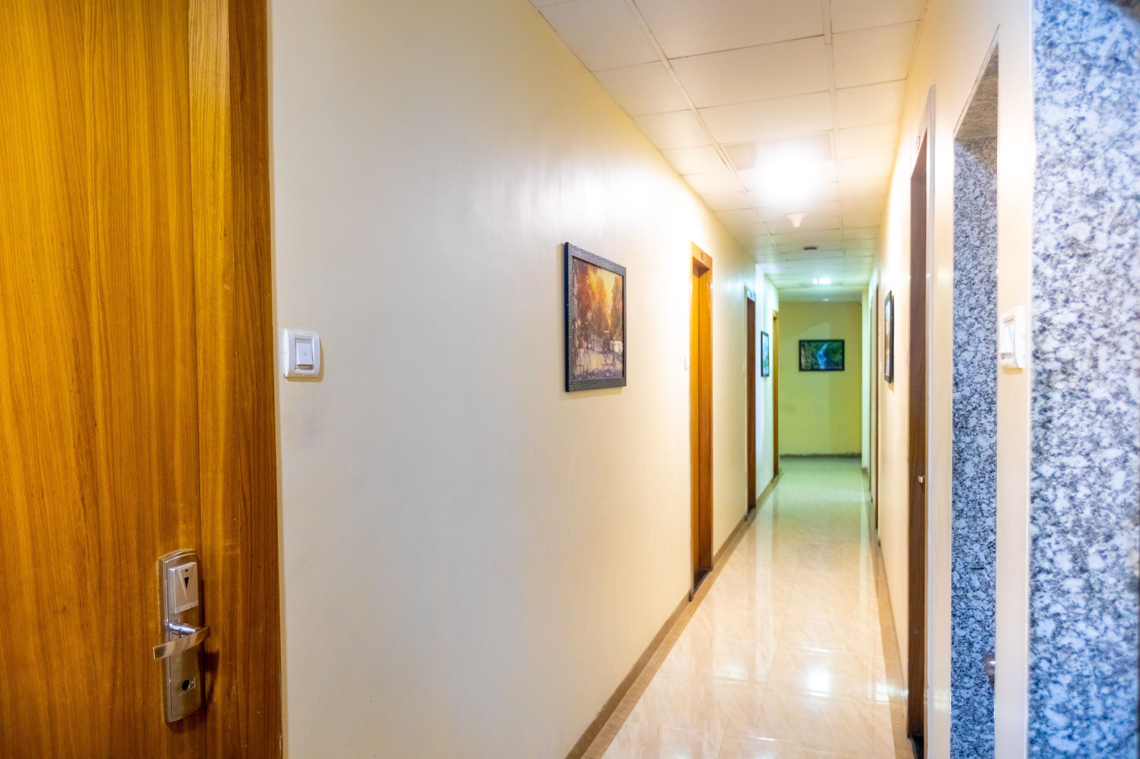A hotel hallway with a wooden door on the left, a marble wall on the right, and a light-colored floor.