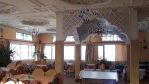 Une salle à manger spacieuse avec de hauts plafonds, des motifs décoratifs élaborés et des lustres. Les tables sont dressées avec des nappes blanches et des chaises.