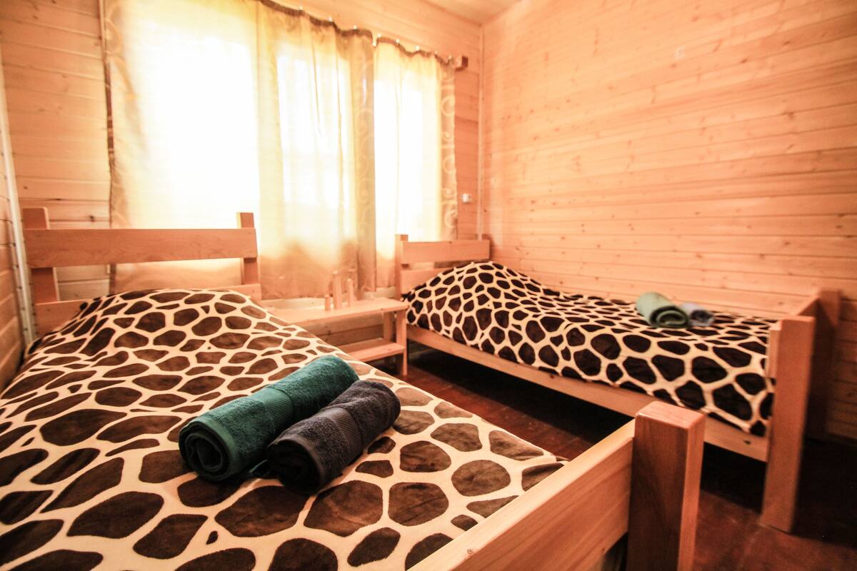 A wooden cabin room with two beds, each with a giraffe-patterned bedspread, a wooden bench, and a window with curtains.