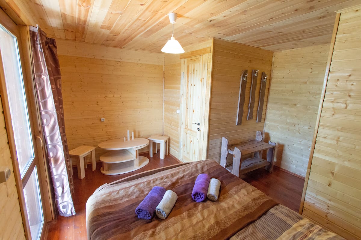A wooden cabin room with a bed, a small table, a bench, and skis leaning against the wall.