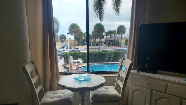 Ein Zimmer mit einem runden Tisch und zwei gepolsterten Sesseln, einem Fernseher auf einem Schrank sowie Blick auf einen Swimmingpool und Palmen.