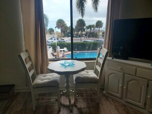 Ein Zimmer mit einem runden Tisch und zwei gepolsterten Sesseln, einem Fernseher auf einem Schrank sowie Blick auf einen Swimmingpool und Palmen.