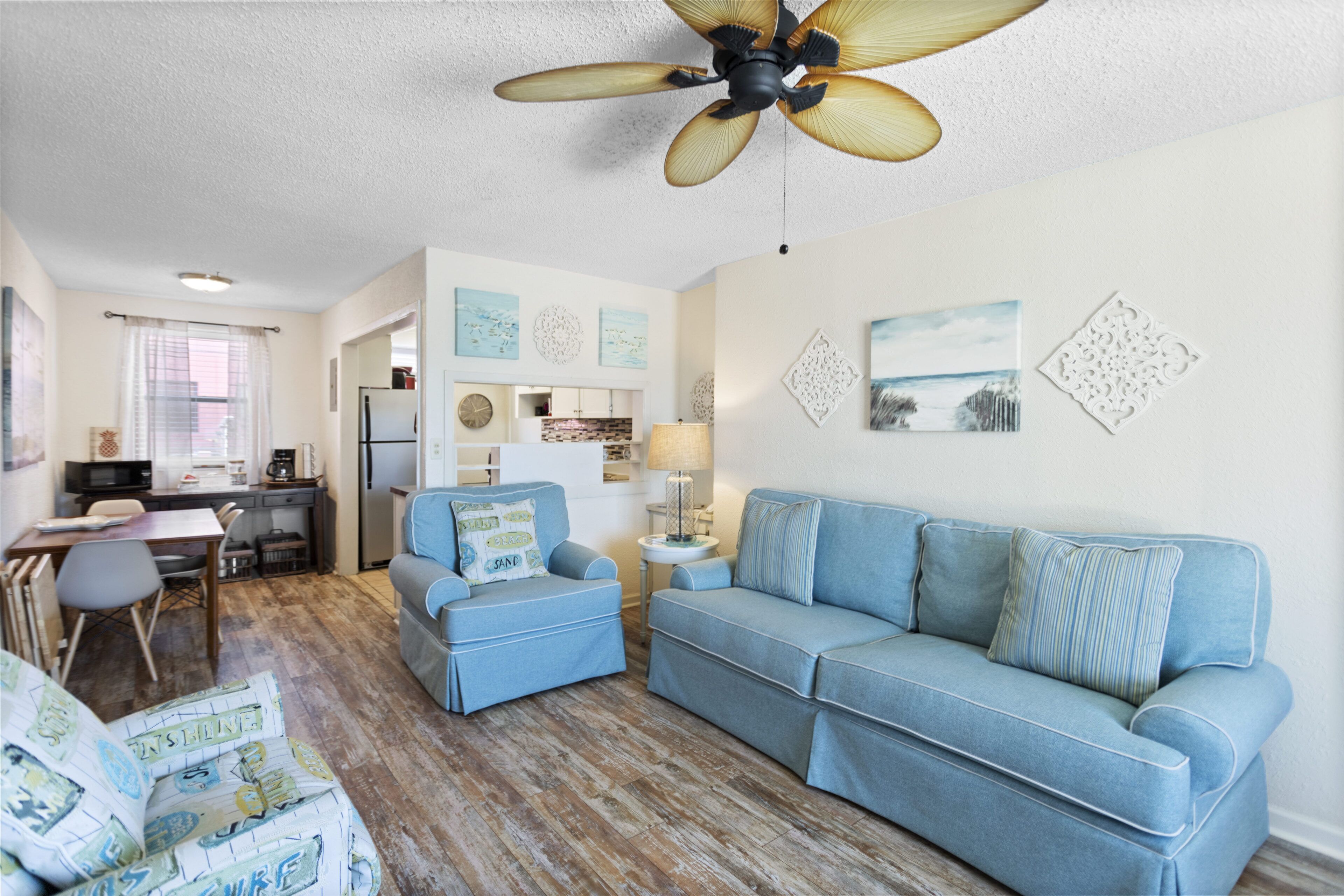 A living room with a blue sofa, a blue armchair, a wooden floor, a ceiling fan, and a kitchen area in the background.