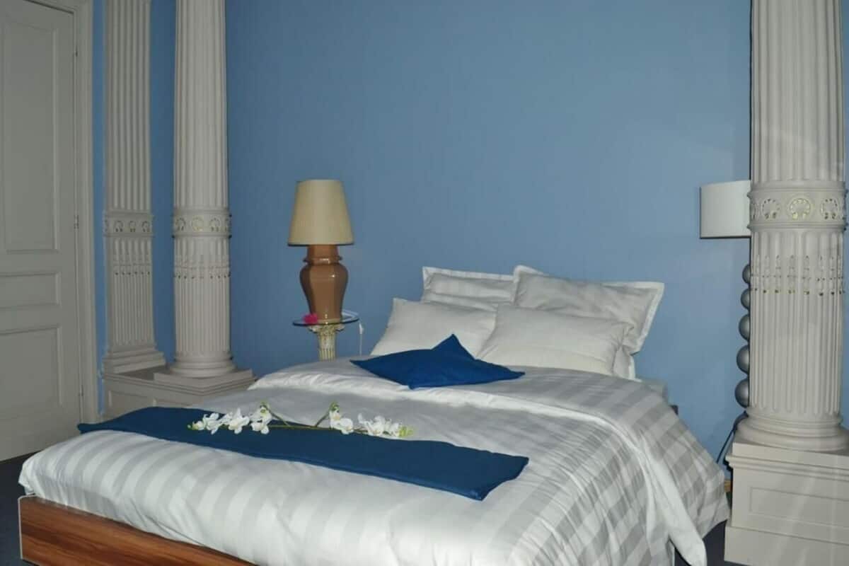 Ein Hotelzimmer mit einem großen Bett, weißen Leinentüchern, blauen Kissen, einer Lampe und dekorativen Säulen.