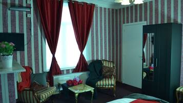 Habitación de hotel con papel tapiz a rayas, una cama, una silla, una mesita, un radiador, un espejo y una ventana con cortinas rojas.
