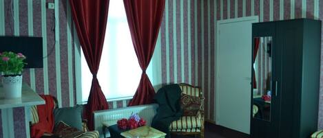Habitación de hotel con papel tapiz a rayas, una cama, una silla, una mesita, un radiador, un espejo y una ventana con cortinas rojas.