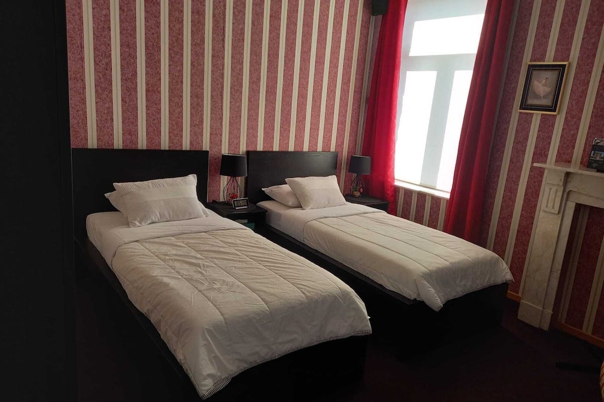 Ein Hotelzimmer mit zwei Einzelbetten, gestreifter Tapete, einem roten Vorhang, einem gerahmten Bild an der Wand und einem Kamin.