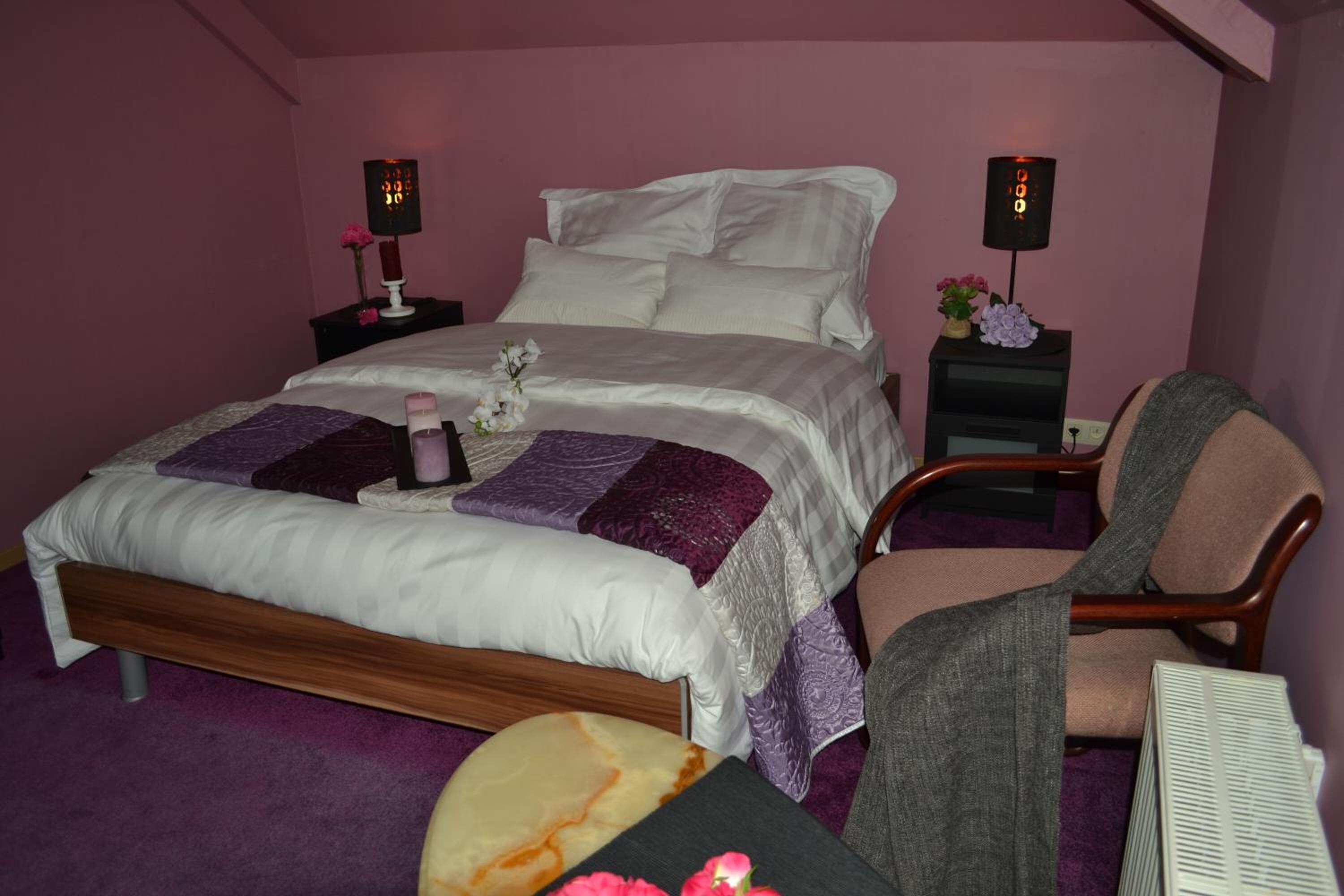 A neatly made bed with a white comforter and purple accents, a wooden chair, and two bedside tables with lamps.