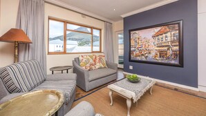 A living room with a sofa, armchair, and a small table with a bowl. A large window provides a view of buildings and mountains. - Vibrant and Safe 2 Bedroom Apartment (Cape Town)
