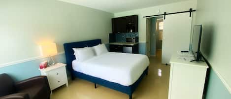 A hotel room with a bed, a nightstand, a desk, a chair, a TV, and a kitchenette.