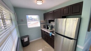 A kitchen with dark cabinets, a stainless steel refrigerator, a microwave, and a window with blinds. - Honu Cove (Age 16+) (Deerfield Beach)