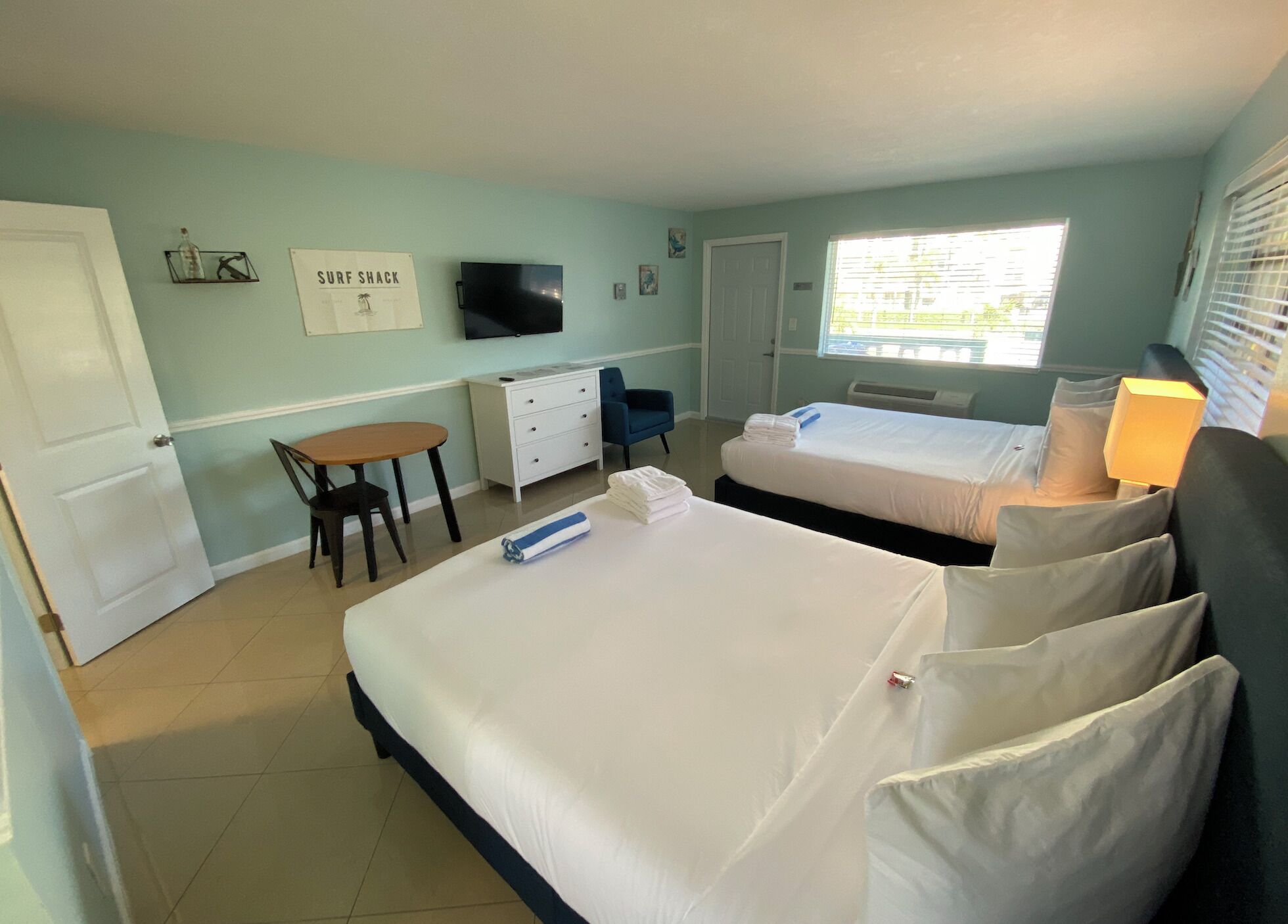 A hotel room with two beds, a desk, a chair, a TV, and a window with blinds.