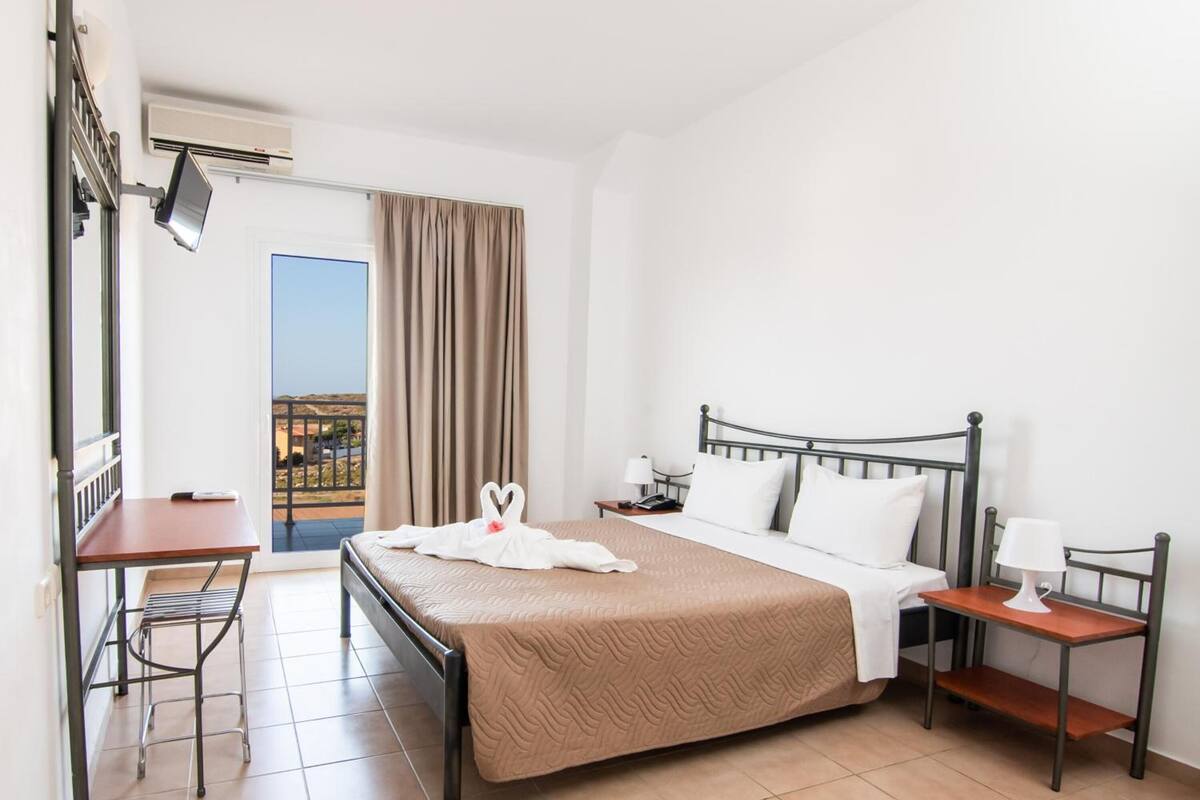 A hotel room with a bed, a desk, a chair, a nightstand, a lamp, and a view of the outdoors.