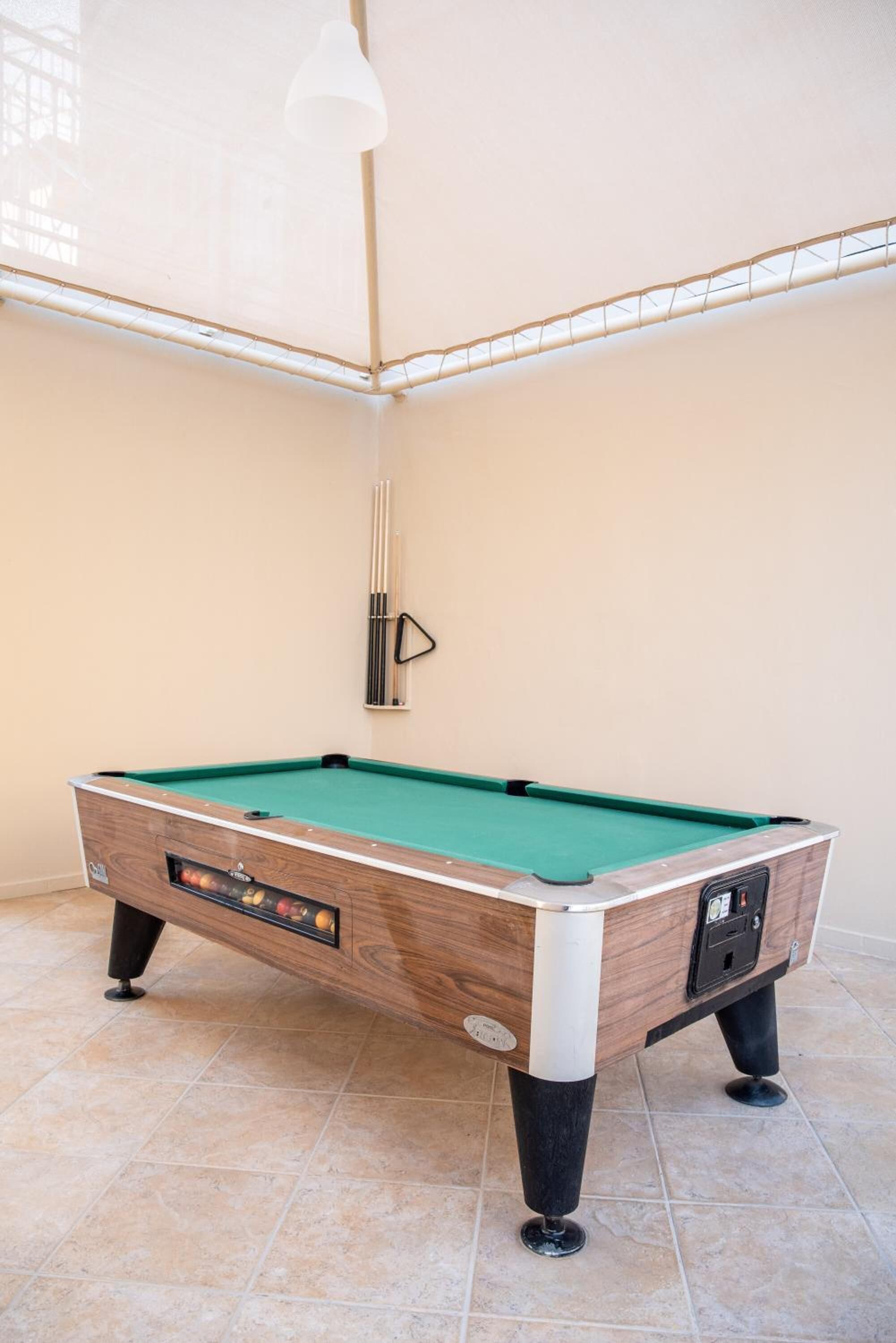 A pool table with a wooden finish and a green felt surface, located in a room with tiled flooring and a high ceiling.