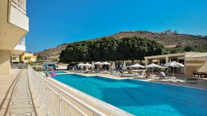 Une piscine dotée de chaises longues et de parasols, entourée d’une balustrade blanche et offrant une vue sur une colline.
