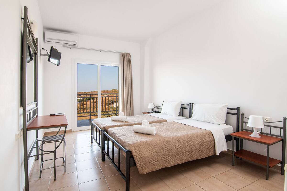 A hotel room with two beds, a desk, a chair, a TV, and a balcony with a view.