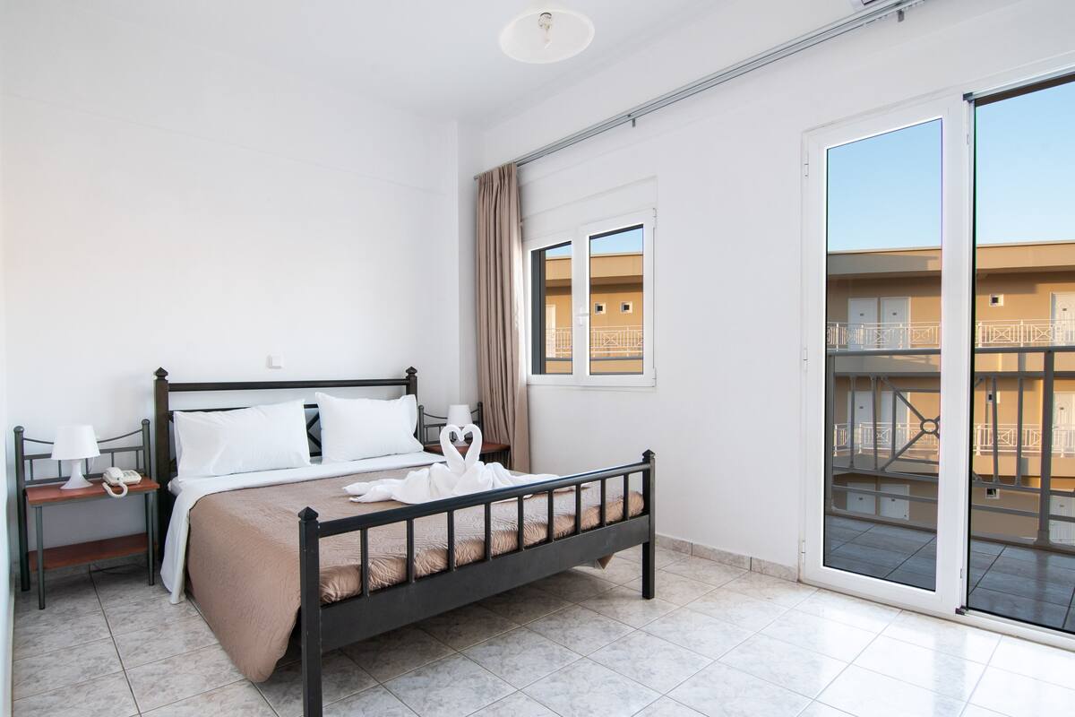 A hotel room with a bed, a nightstand, a lamp, a window, and a view of the outside balcony.