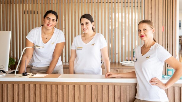Tres recepcionistas con uniformes blancos de pie detrás de un mostrador de recepción en una oficina moderna con paneles de madera y un monitor de ordenador.
