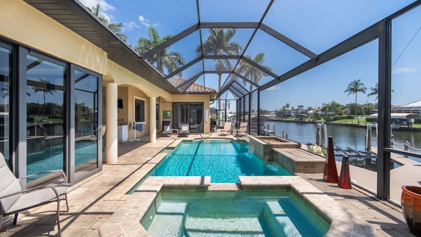 A pool area with clear views of a lake, surrounded by glass walls and palm trees. - 4BR gulf access villa w/heated pool & hot tub! Villa Francesca - SWFL Rentals (Cape Coral)