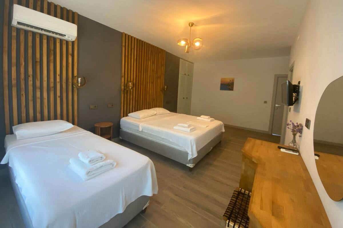 Hotelkamer met twee bedden, een wand van houten latten, een airconditioningapparaat, een kleine ronde tafel, een houten bankje en een aan de muur bevestigde televisie.