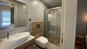 Moderni kylpyhuone, jossa on suuri peili, valkoinen pesuallas, wc ja lasiseinällä varustettu suihku.