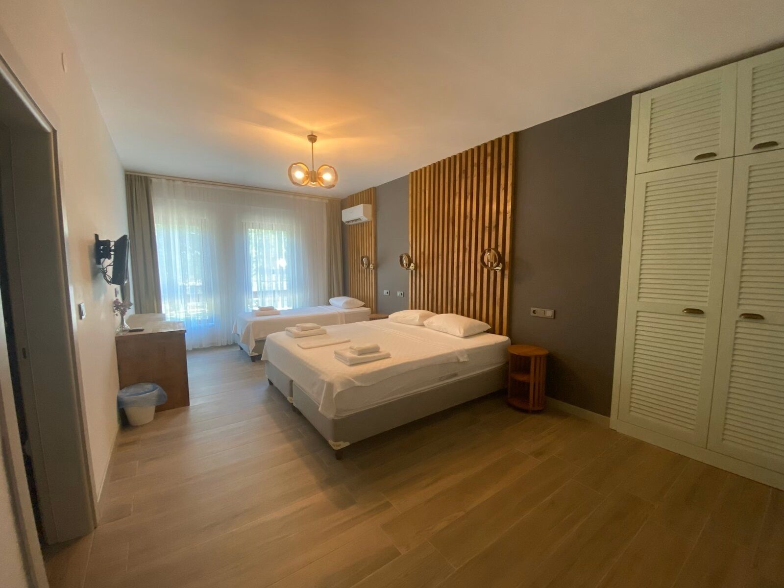 A modern hotel room with two beds, a wooden floor, a small bedside table, a mirror, and a wardrobe.