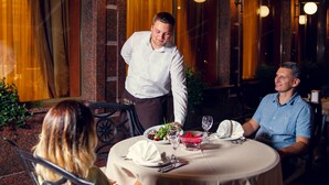 A restaurant with a round table set for two, a waiter serving a meal, and a dining area with wooden accents and hanging lights. - Solva Resort & SPA (Poliana)
