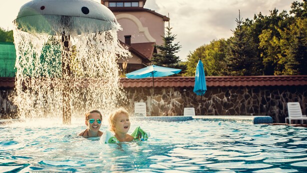A swimming pool with two people in it, a water fountain, a blue umbrella, and a building in the background. - Solva Resort & SPA (Poliana)