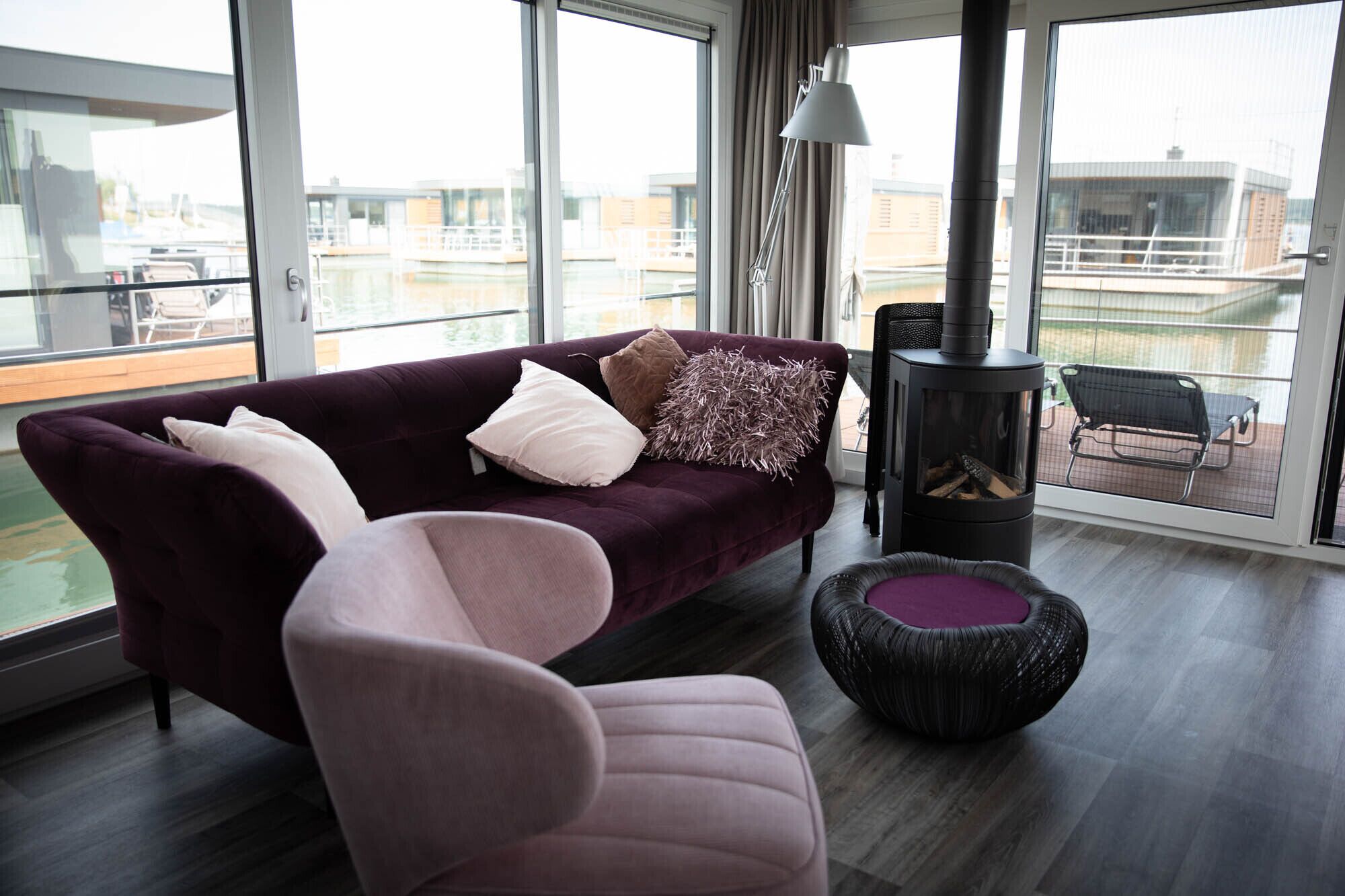 Een moderne woonkamer met een paarse bank, een rond hockertje en een houtkachel.