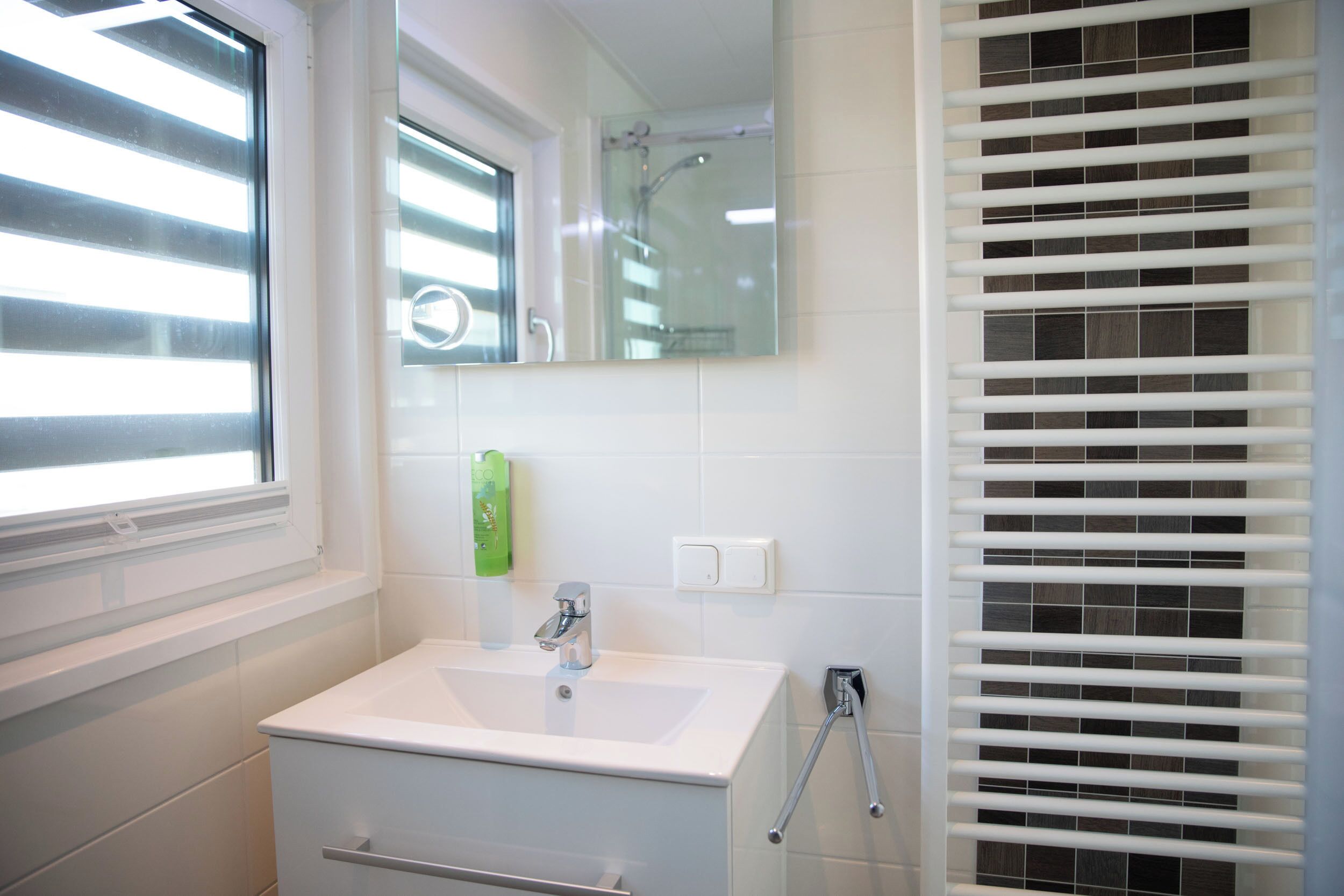 Ein Badezimmer mit Waschbecken, einem Spiegel, einem Fenster mit Jalousien und einer Duschkabine.