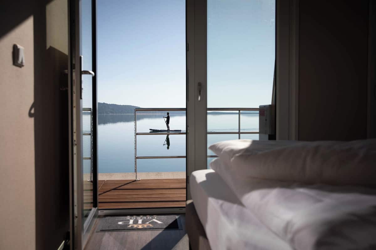 A room with a bed and a view of a calm lake through a glass door.