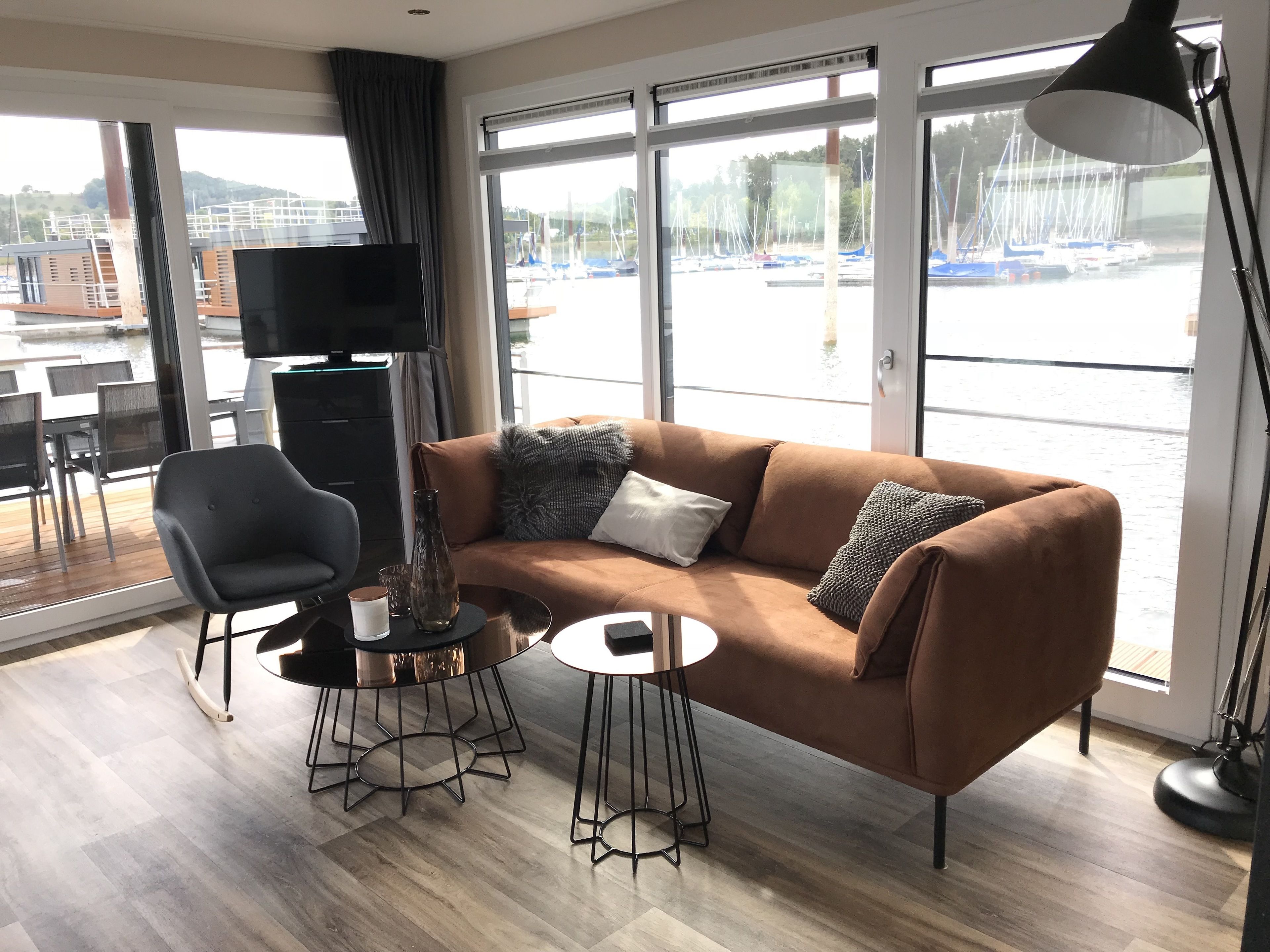 Een moderne woonkamer met een bruine bank, een zwarte fauteuil en een kleine ronde tafel. Een televisie is aan de muur bevestigd, en er is een groot raam met uitzicht op boten en een jachthaven.