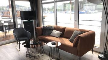 Een moderne woonkamer met een bruine bank, een zwarte fauteuil en een kleine ronde tafel. Een televisie is aan de muur bevestigd, en er is een groot raam met uitzicht op boten en een jachthaven.