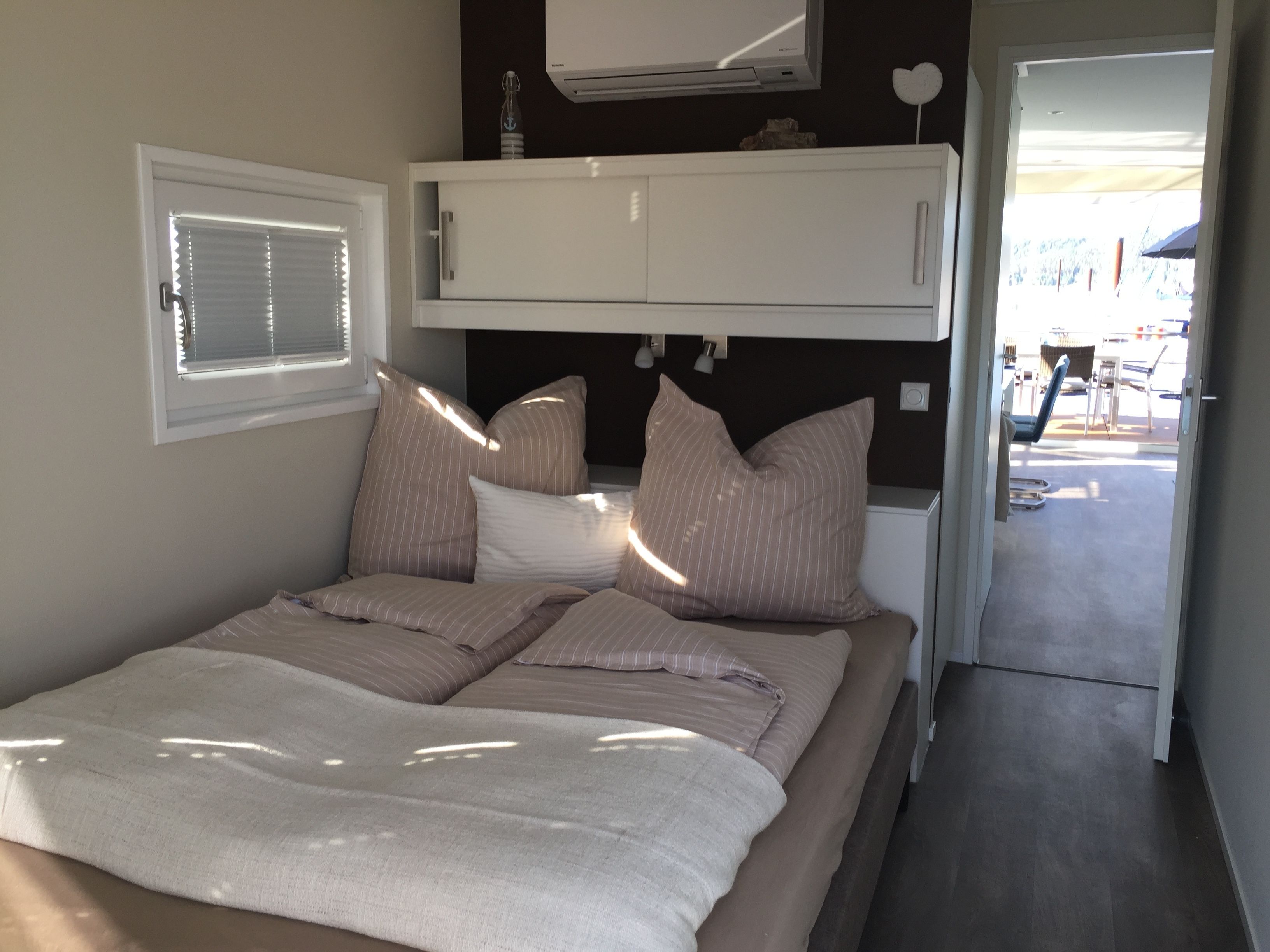 A bedroom with a bed, pillows, an air conditioning unit, a window with blinds, and a view of the outside.