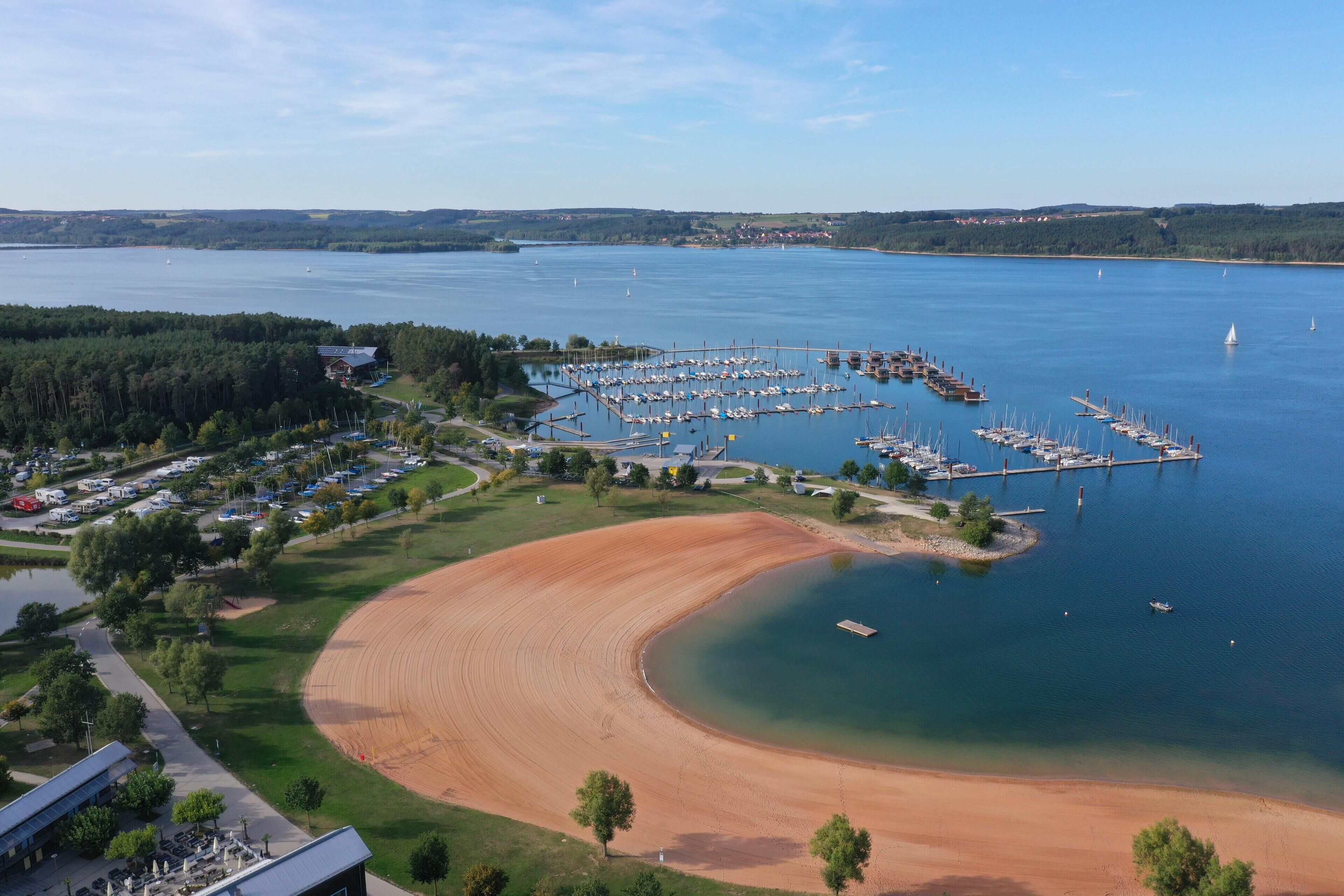 Vue aérienne d’une zone côtière comprenant un port de plaisance, une plage de sable et une végétation luxuriante environnante.