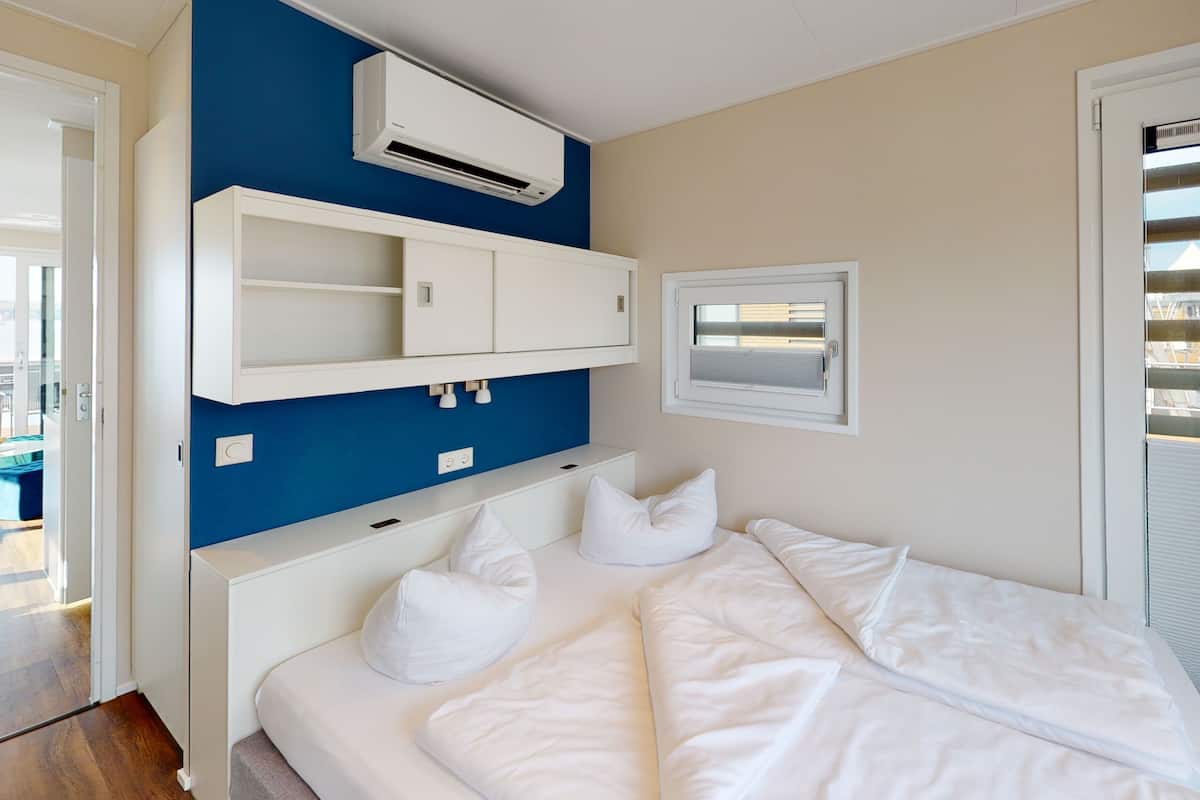 A bedroom with a bed, white pillows, a blue accent wall, an air conditioning unit, a window with blinds, and a view of buildings outside.