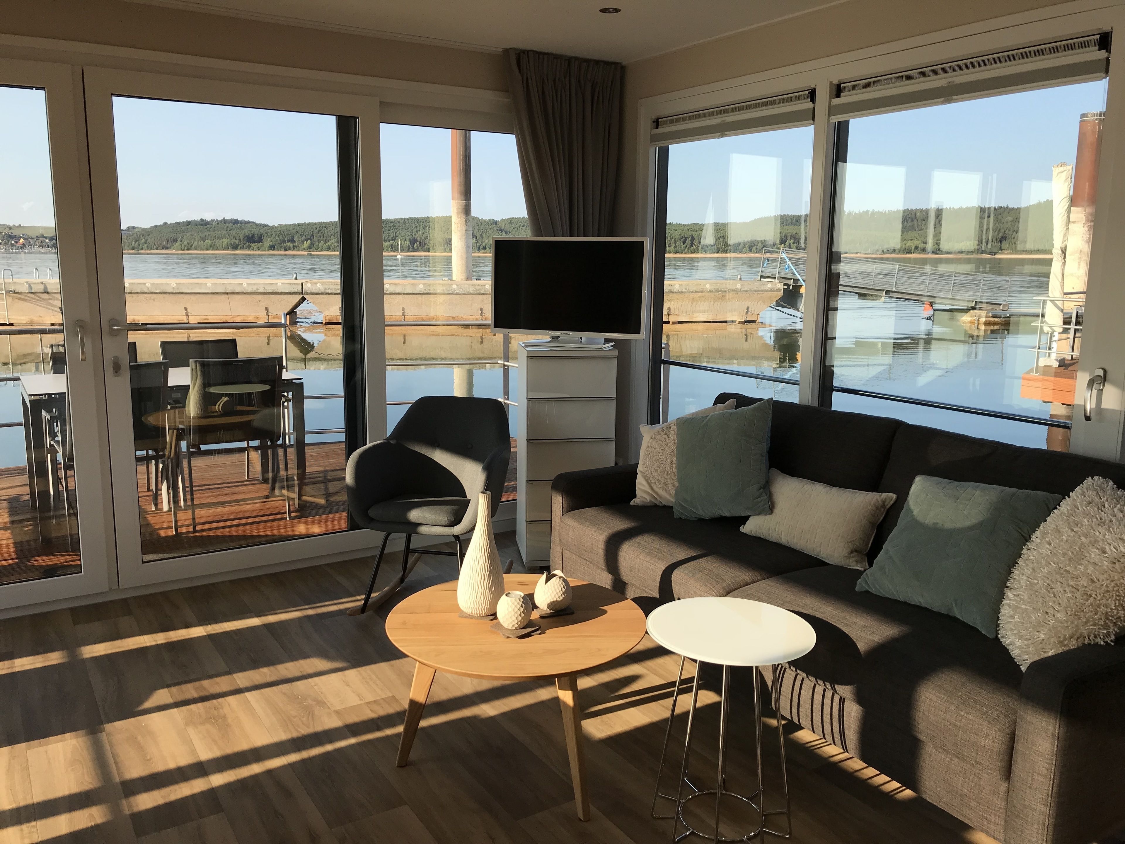 A modern living room with a sofa, coffee table, and TV, overlooking a beach and water body.